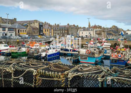 De nombreux petits bateaux de pêche sont amarrés en sécurité aux quais du port de Fraserburgh, avec des bâtiments en pierre bordant le front de mer sous un ciel nuageux Banque D'Images