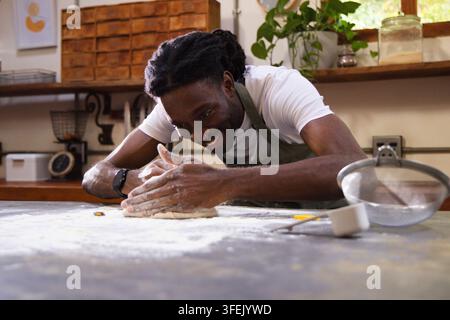 Homme africain portant un tablier vert, penché sur un plan de travail de cuisine fariné en train de pétrir une boule de pâte avec une passoire Banque D'Images