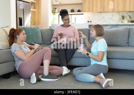 Diverses amies féminines en conversation, vêtues de vêtements de sport, sirotant des gobelets transparents avec des pailles dans un appartement Banque D'Images