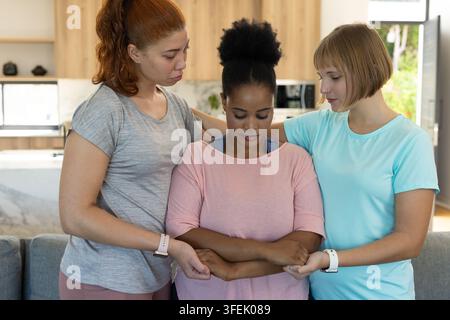 Trois femmes réconfortant une femme centrale debout dans la cuisine près d'un canapé gris avec des bracelets de fitness Banque D'Images