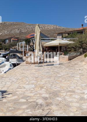 Pirovac, Croatie, 16 août 2026 Terrasse en bord de mer ensoleillée avec pavage en pierre, parasols beiges, bateaux amarrés, bâtiments rustiques et arrière-plan montagneux rocheux Banque D'Images