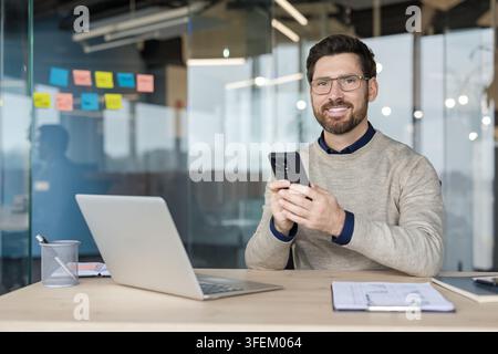 Homme d'affaires professionnel portant des lunettes et un pull smart-casual sourit à l'objectif tout en envoyant des messages sur son smartphone à un bureau lumineux équipé d'un ordinateur portable, exprimant productivité et connectivité moderne Banque D'Images