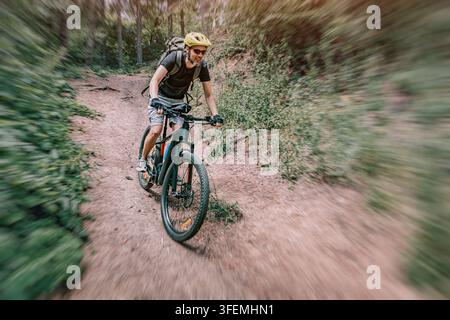 Jeune homme profitant d'une aventure en VTT, portant un casque et un sac à dos, descendant rapidement une pente en forêt sur un sentier poussiéreux Banque D'Images