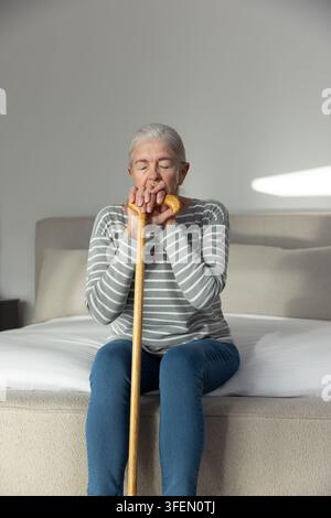 Sénior femme assise au bord du lit tenant une canne en bois, les yeux fermés, portant un jean Banque D'Images