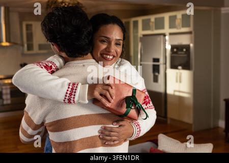 Couple afro-américain s'embrassant dans une cuisine chaleureuse, femme tenant un cadeau emballé derrière le dos de son partenaire Banque D'Images