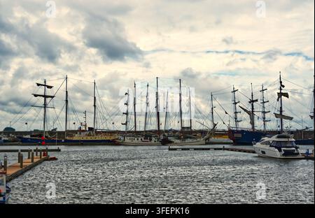 Grands voiliers historiques à côté d'un yacht de luxe amarré dans un port animé, mettant en valeur des embarcations nautiques sous un ciel nuageux Banque D'Images