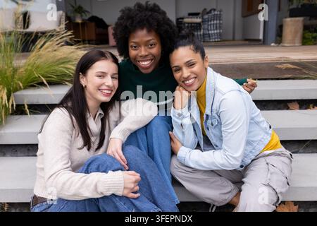 Différentes amies féminines assises sur des marches en béton au porche avec des bacs à fleurs, portant des pulls et des jeans Banque D'Images