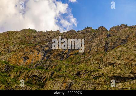 La paroi rocheuse des montagnes Rocheuses met en valeur des textures rugueuses et une végétation verte. Une lumière vive illumine le terrain escarpé sous un ciel clair parsemé de nuages. Banque D'Images