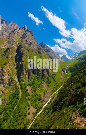 Vue en contre-plongée capturant des pentes montagneuses vertes déchiquetées et des sommets lointains enneigés. Une rivière sinueuse cascade le long du flanc rocheux de la montagne sous un ciel clair Banque D'Images
