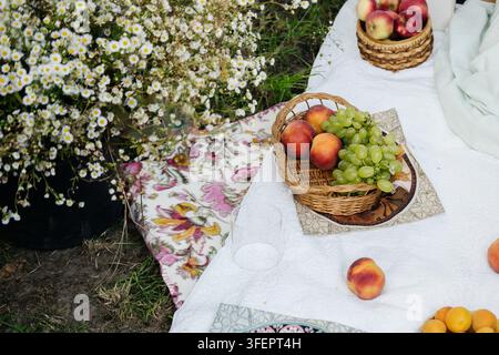 Nappe de pique-nique blanche étalée sur l'herbe avec des paniers en osier remplis de pêches, de raisins et de pommes, un tissu fleuri et des abricots éparpillés, à côté d'un seau de Banque D'Images