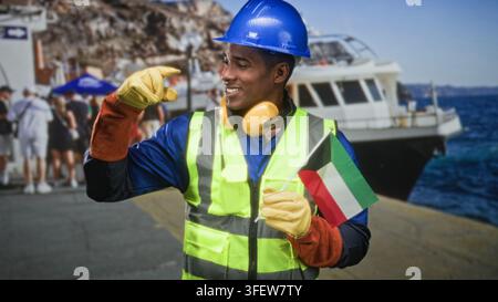Homme tenant le drapeau du Koweït dans une main gantée et pointant avec l'autre main vers le quai près du bateau, portant un casque de sécurité et un gilet de haute visibilité ; fierté devoir. Banque D'Images