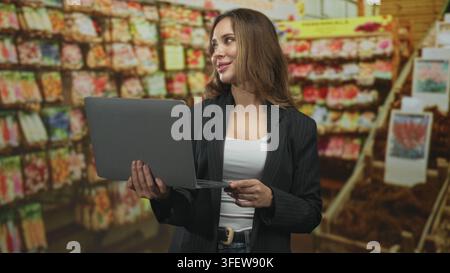 Une femme tient un ordinateur portable et scanne les étagères des paquets de semences tout en vérifiant les niveaux de stock dans l'immeuble ; planification de la vente au détail en toute confiance. Banque D'Images