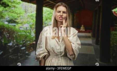 Femme blonde touchant sa joue avec ses doigts pour une douleur dentaire à l'intérieur d'un couloir d'un bâtiment couvert bordé de plantes et de colonnes ; inconfort. Banque D'Images