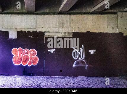 Un mur de viaduc en béton arborant des graffitis peints au spray, dont une pièce en lettres bulle orange vif à gauche et un personnage de dessin animé blanc stylisé Banque D'Images