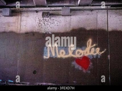 Une œuvre d'art de rue peinte en spray sur un mur de béton érodé, représentant le nom "Melodie" en caractères clairs à côté d'un cœur rouge plein. Banque D'Images
