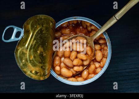 Haricots cuits en conserve dans une sauce tomate avec cuillère, vue de dessus, gros plan. Boîte de conserve ouverte remplie de délicieux haricots blancs cuits dans une riche sauce tomate avec un o Banque D'Images