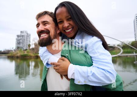Heureux couple multiracial piggyback et souriant à l'extérieur. Banque D'Images