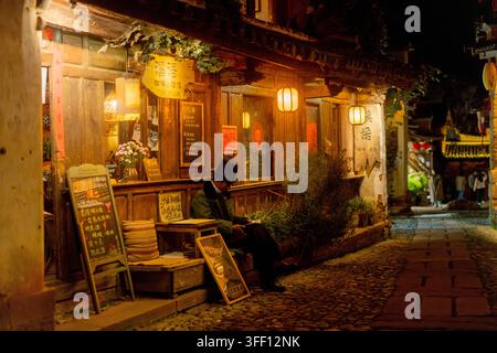 Homme en haut-de-forme assis seul devant un café la nuit, vieille ville de Shaxi, Yunnan, Chine Banque D'Images