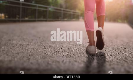 Un plan rapproché, pris de bas, d'une femme marchant sur un sentier extérieur, portant un legging rose et des baskets blanches, capturant un sens de style de vie actif Banque D'Images
