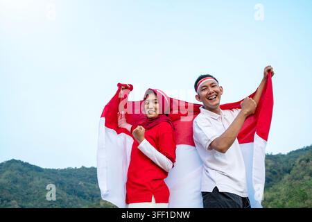 Un homme et une femme indonésiens portant le hijab tenant ensemble le drapeau indonésien lors d'une célébration du Jour de l'Indépendance avec un fond de montagne. Hari Kemerdekaan Banque D'Images