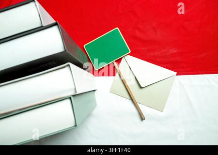 Une pile de livres, une enveloppe et un panneau en bois vert vierge sur le drapeau rouge et blanc de l'Indonésie. Idéal pour les fêtes éducatives, nationales ou patri Banque D'Images