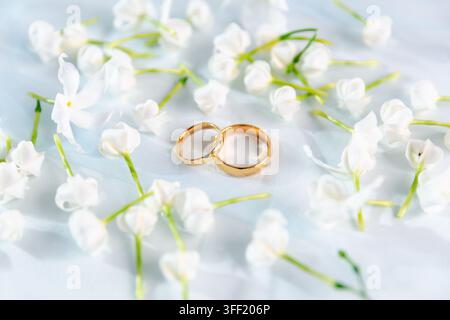 Deux alliances en or entourées de délicates fleurs blanches sur un tissu blanc doux, créant une scène de mariage élégante et romantique symbolisant l'amour, le mariage Banque D'Images