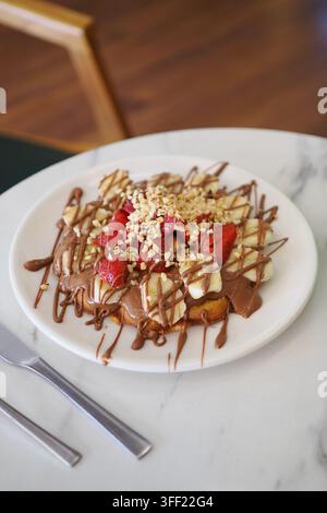 Gaufre servie sur une assiette blanche, garnie de bananes, de fraises, de noix et d'une sauce au chocolat Banque D'Images