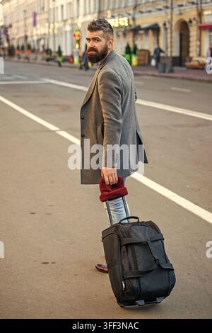 Homme d'affaires en voyage sur le chemin. Voyageur touristique barbu marchant dans la rue de la ville. Touriste avec sac en ville. Homme élégant barbu marchant et traversant Banque D'Images