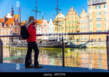 Touriste en veste rouge avec sac à dos promenades le long du front de mer de Gdansk. Derrière elle se trouvent des bâtiments historiques avec des façades vibrantes et une voile traditionnelle Banque D'Images
