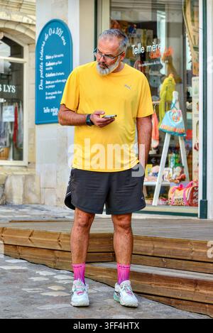Homme d'âge moyen barbu portant un T-shirt jaune, un short, des chaussettes roses et des baskets tenant un smartphone devant un magasin - Loches, Indre-et-Loire (37), France. Banque D'Images