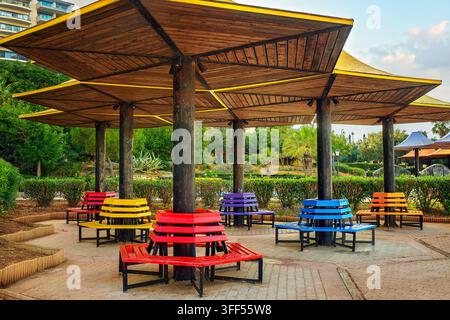 Des bancs circulaires lumineux peints aux couleurs de l'arc-en-ciel sous des auvents en bois en forme de parapluie dans un parc public. Banque D'Images