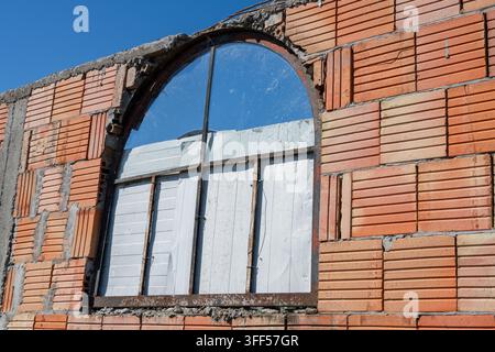un mur extérieur inachevé en briques de terre cuite creuses, avec un cadre de fenêtre métallique rouillé et arqué partiellement condamné par des planches blanches Banque D'Images