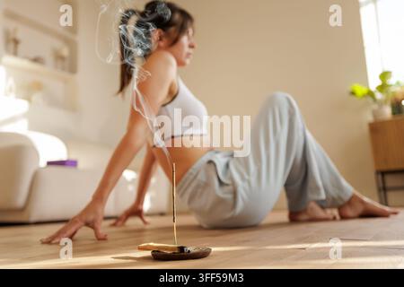 De l'encens naturel brûle sur un porte-encens en bois tandis qu'une jeune femme se détend sur le sol à la maison, suggérant l'aromathérapie, une routine apaisante et une mentalité consciente Banque D'Images