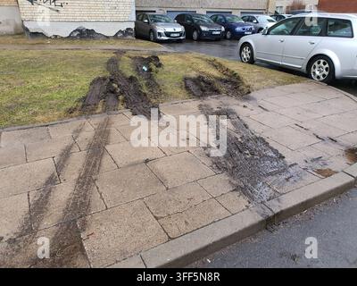 Des traces de pneus profondément enfouées dans la boue marquent un trottoir pavé et la zone herbeuse adjacente dans un cadre résidentiel urbain. La boue brun foncé crée des ornières proéminentes Banque D'Images