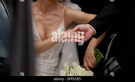 Close up des mains jointes d'un couple marié près d'une voiture noire brillante. Une lumière naturelle douce met en valeur la robe en dentelle et les fleurs blanches. Ambiance romantique et élégante wit Banque D'Images