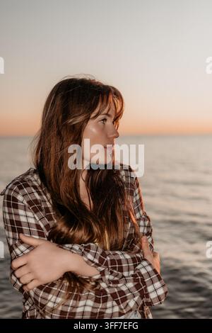 Femme au coucher du soleil sur l'océan, debout au bord de l'eau, portant une chemise à carreaux et regardant pensivement au loin pendant l'heure dorée Banque D'Images