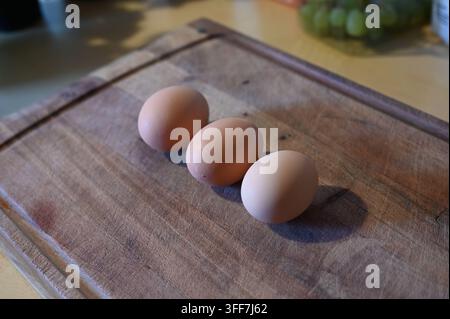 Trois œufs de poule bruns sur une planche à découper rustique en bois dans une cuisine. Banque D'Images