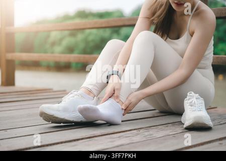 Femme fitness courant ressentant une douleur à la cheville et à la jambe pendant l'exercice, assise sur un pont en bois en plein air, tenant son pied blessé avec les mains après une entorse ou Banque D'Images
