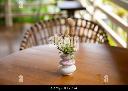 Un petit vase en céramique avec arrangement floral, table en bois, herbe sèche et fleur blanche sur la table de patio avec chaise en rotin en arrière-plan, décoration d'intérieur rustique et calme Banque D'Images
