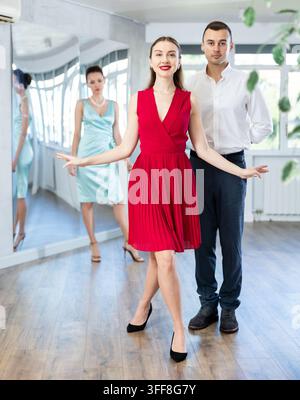 Jeune femme souriante en robe rouge et homme en tenue formelle répétant la révérence et la courbette dans une salle de danse sous la direction d'une enseignante féminine intéressée. T Banque D'Images