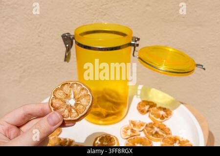 Main tenant une tranche de citron séchée avec un bocal et une assiette de citrons séchés Banque D'Images