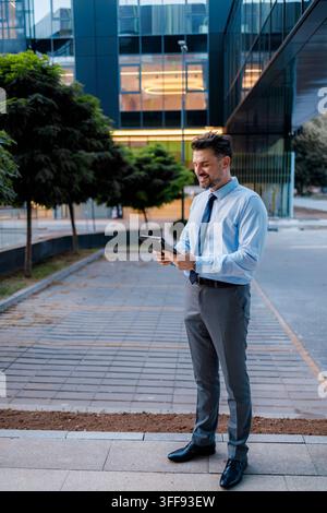 Un homme en tenue formelle se tient sur un chemin, utilisant une tablette. Il est à l'extérieur d'un grand bâtiment moderne aux murs de verre. Banque D'Images