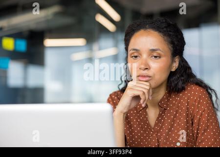 Femme professionnelle aux cheveux bouclés pensant sérieusement, concentrée sur son travail, assise à son bureau dans un bureau moderne, utilisant un ordinateur portable, représentant le dévouement et la résolution de problèmes dans les affaires Banque D'Images