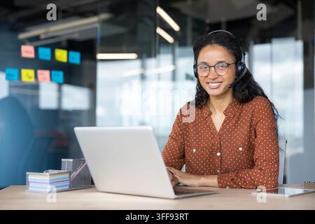 Agent de service client heureuse portant un casque et des lunettes, souriant tout en étant assise à son bureau et travaillant sur un ordinateur portable dans un environnement de bureau moderne Banque D'Images