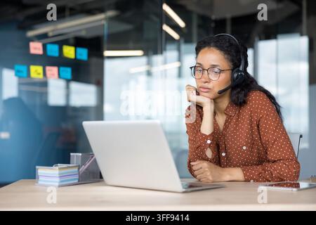 Agent de centre d'appel portant un casque et des lunettes, paraissant fatigué et ennuyé tout en regardant un écran d'ordinateur, fournissant un support client ou participant à une réunion d'affaires à distance dans un environnement de bureau Banque D'Images