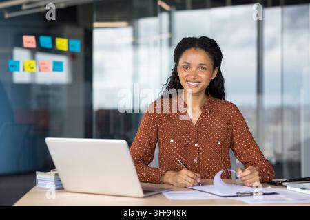 Jeune femme professionnelle souriant à l'appareil photo tout en travaillant sur son ordinateur portable et en prenant des notes à un bureau dans un espace de travail d'entreprise contemporain, incarnant la productivité et le succès Banque D'Images