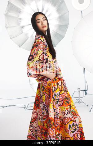 Portrait de mode en studio d'une jeune femme asiatique portant une robe maxi fluide orange et brune à grand motif floral Paisley. Banque D'Images