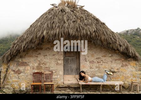Une jeune femme tatouée se détend sur un banc en bois devant une cabane en pierre rustique avec un toit de chaume traditionnel, défilant sur son smartphone entourée de Banque D'Images