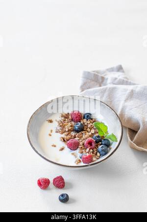 Granola avec yaourt, framboise et myrtilles dans un bol sur un fond blanc avec serviette et lumière naturelle. Concept de petit-déjeuner sain à base de produits laitiers Banque D'Images