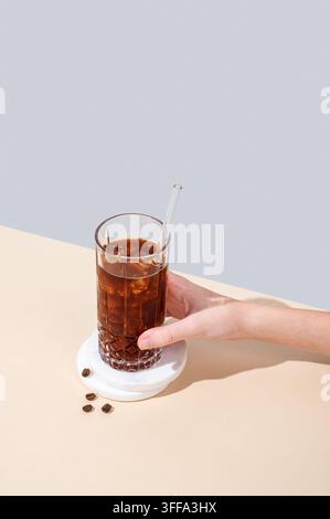 Main tenant un verre de café froid avec glace et paille sur un fond jaune et bleu avec des grains de café et de l'ombre. Boisson moderne rafraîchissante d'été. Banque D'Images